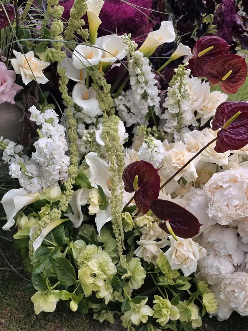 A large floral arrangement with white calla lilies, white hydrangeas, white roses, and dark red anthuriums, accented with green foliage and tall green amaranthus.