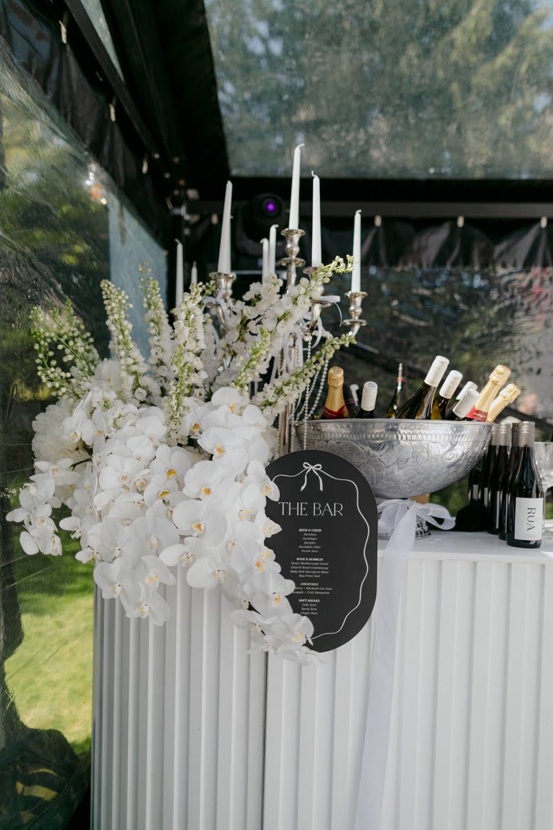 A bar setup with a bouquet of white orchids and flowers, candles, and bottles of champagne.