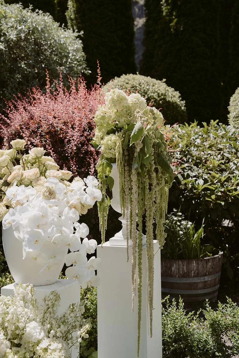 Arrangement of flowers in vases and planters outdoors, with greenery and bushes in the background.