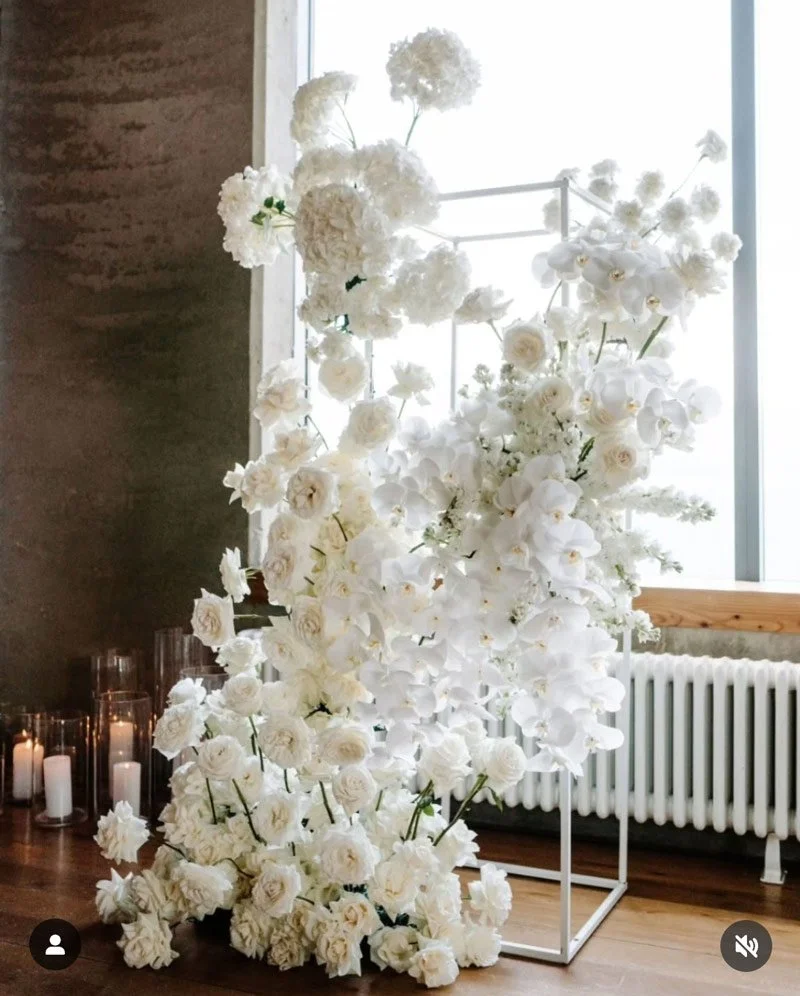Large floral arrangement of white flowers, including roses and orchids, on a stand next to a window with candles on the floor nearby.