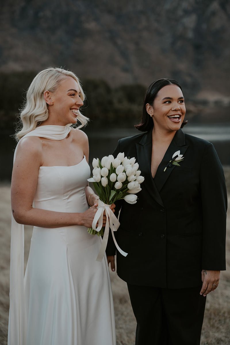Two women, holding a bouquet of white tulips, standing outdoors with a body of water and mountains in the background, smiling and laughing.