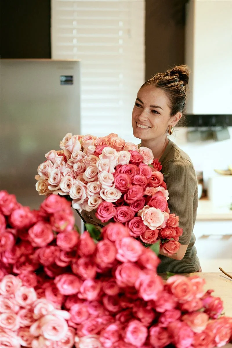 A woman with brown hair in a bun smiling while holding a large bouquet of pink and white roses in a kitchen.