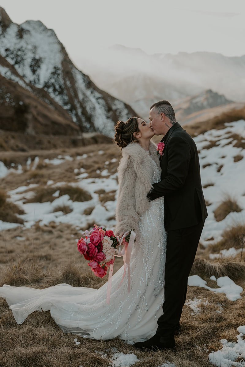 A couple in wedding attire kissing in a mountainous, snowy landscape, with the bride holding a bouquet of pink and white flowers.