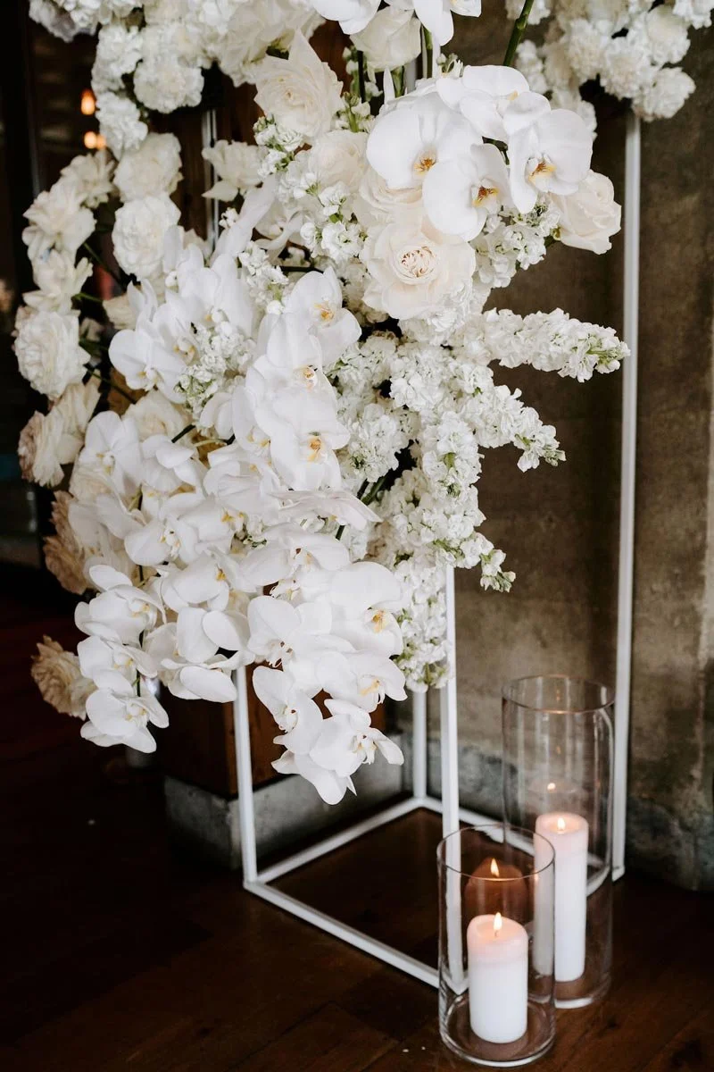 A floral arrangement of white orchids and other white flowers inside a white metal frame, with three candles in glass holders at the base.