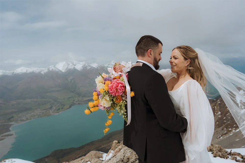 A bride and groom sharing a romantic moment on a mountain top during their wedding, with a lake and snow-capped mountains in the background.