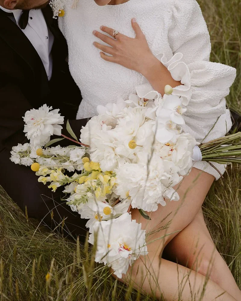 A couple, sitting outdoors in a grassy field. The woman is holding a large bouquet of white flowers.
