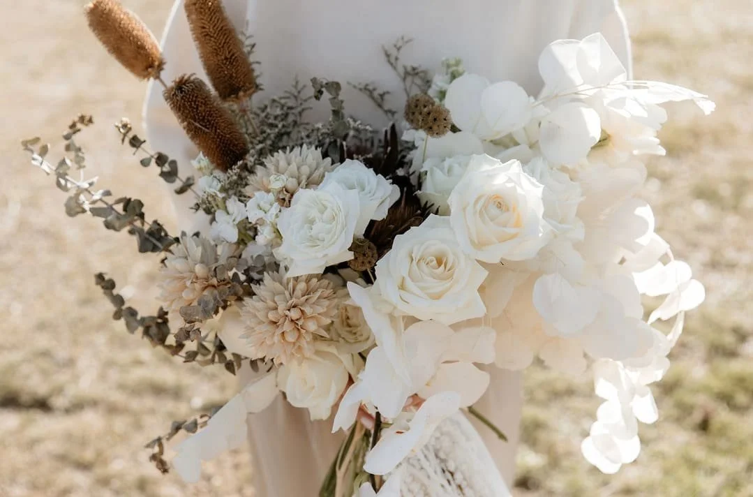 Bouquet of white roses, dried beige flowers, greenery, and beige fuzzy accents, held outdoors with a bright background.