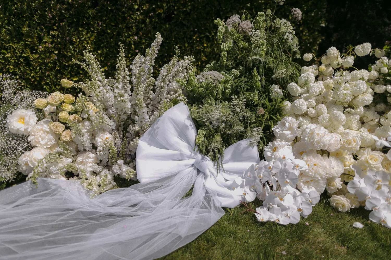 A floral arrangement with white roses, orchids, and other white flowers, decorated with a large white bow and white tulle fabric, placed on a grassy area, likely for a wedding or special event.