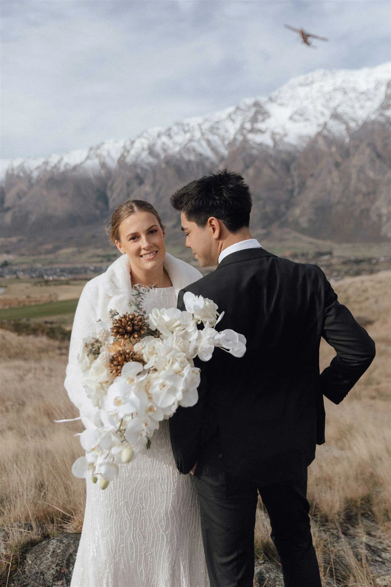 A bride and groom standing outdoors in a field with snow-capped mountains in the background, the bride holding a large bouquet of white flowers and the groom smiling at her.