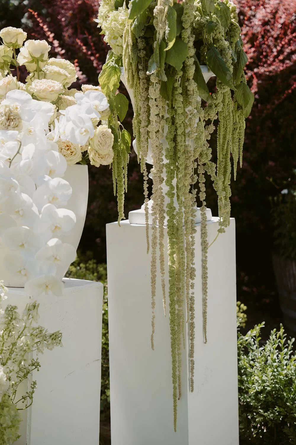 White and cream flowers arranged on a white stand with hanging green sprigs, outdoor garden setting.