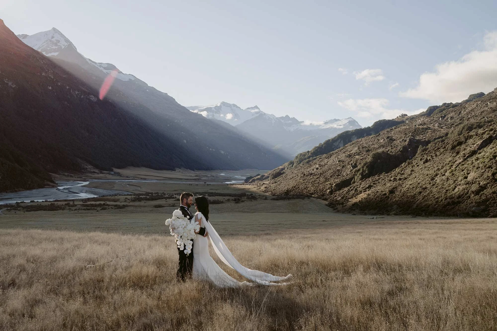 A bride and groom standing in a grassy field with mountains and a river in the background, during sunset or sunrise.