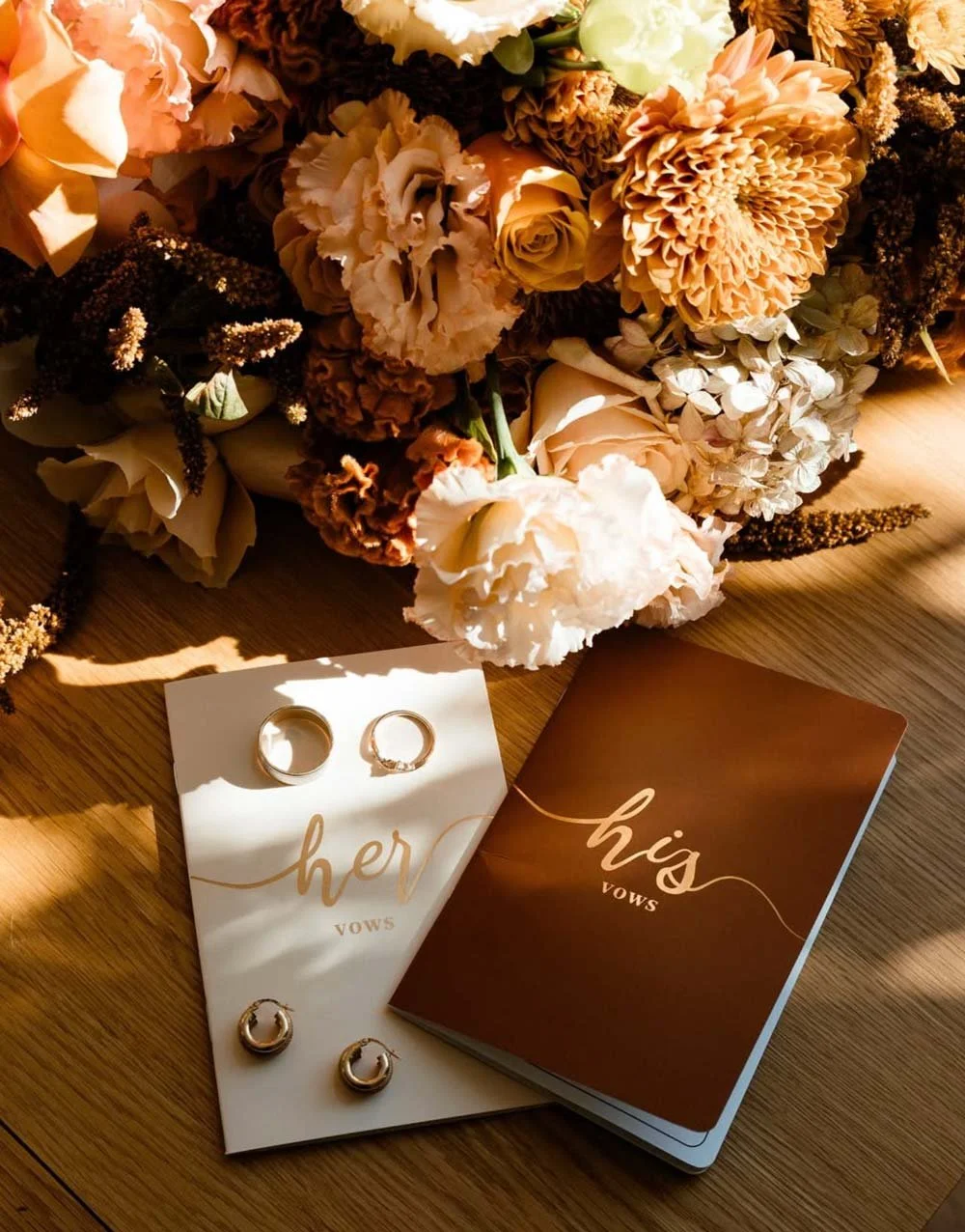 A bouquet of peach, cream, and white flowers, along with wedding rings on vow booklets labeled 'her' and 'his'.