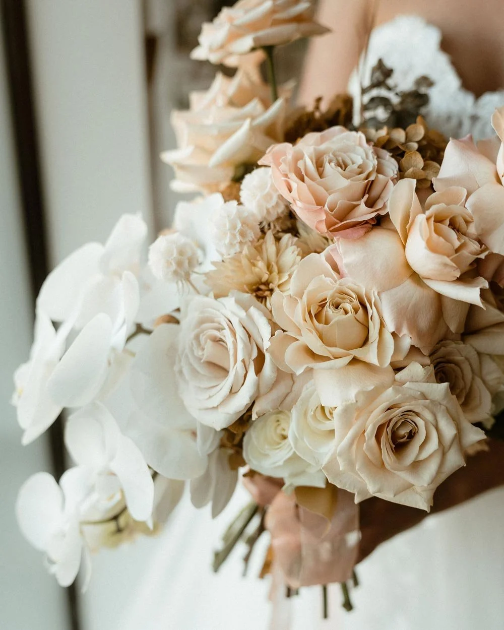 Close-up of a bouquet of cream and blush roses, white orchids, and other flowers, held with a pink ribbon.
