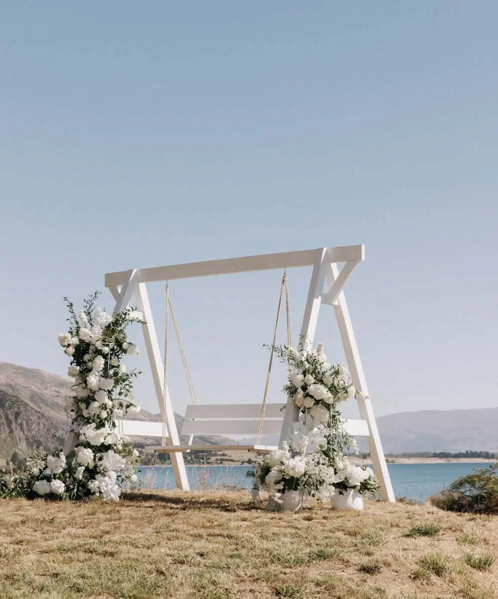 A white wooden swing decorated with white flowers and greenery, situated outdoors near a body of water with mountains in the background.