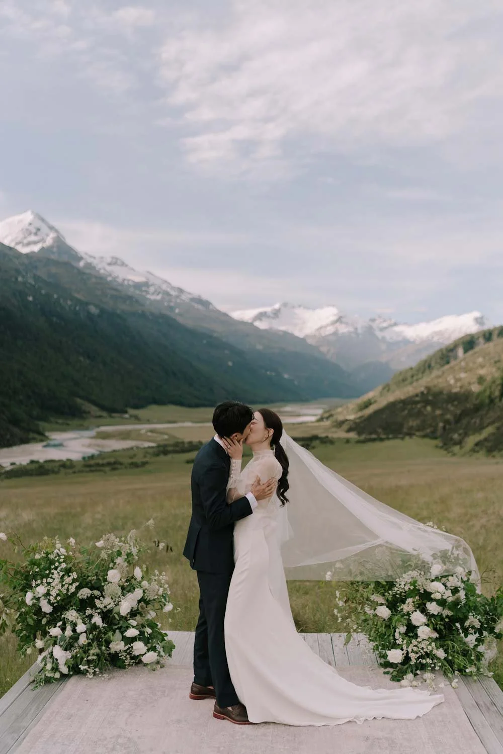 A bride and groom kiss during their outdoor wedding in a mountainous landscape with snow-capped peaks and a river in the background, floral arrangements on a wooden platform.