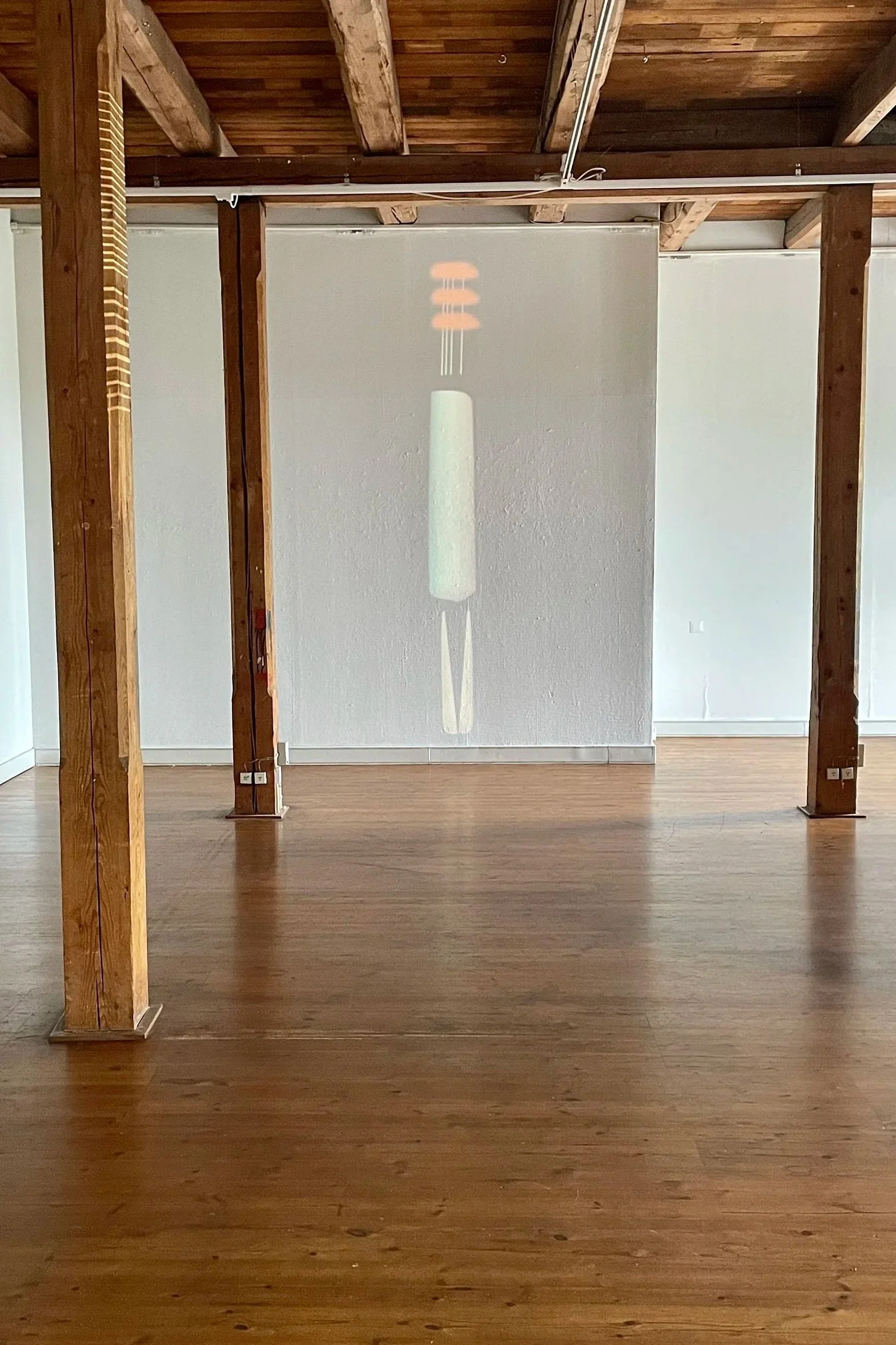 An empty room with wooden floors and exposed wooden beams on the ceiling. There is a wall in the background with a projection of a cylindrical object and some orange shapes above it.