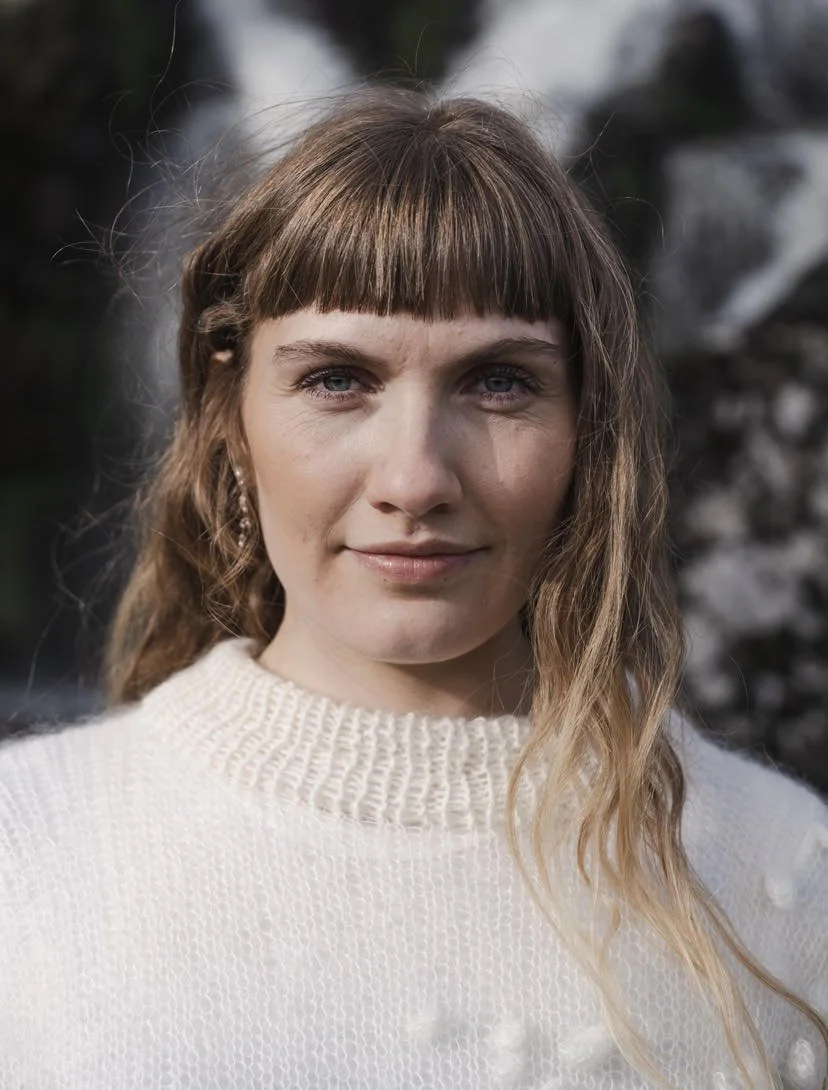 A woman with light skin and brown hair with blonde tips, wearing a cream sweater, standing outdoors with blurred trees in the background.