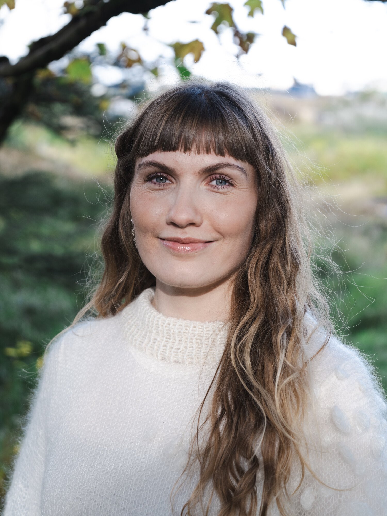 A woman with long wavy hair and bangs, wearing a white sweater outdoors, smiling at the camera.