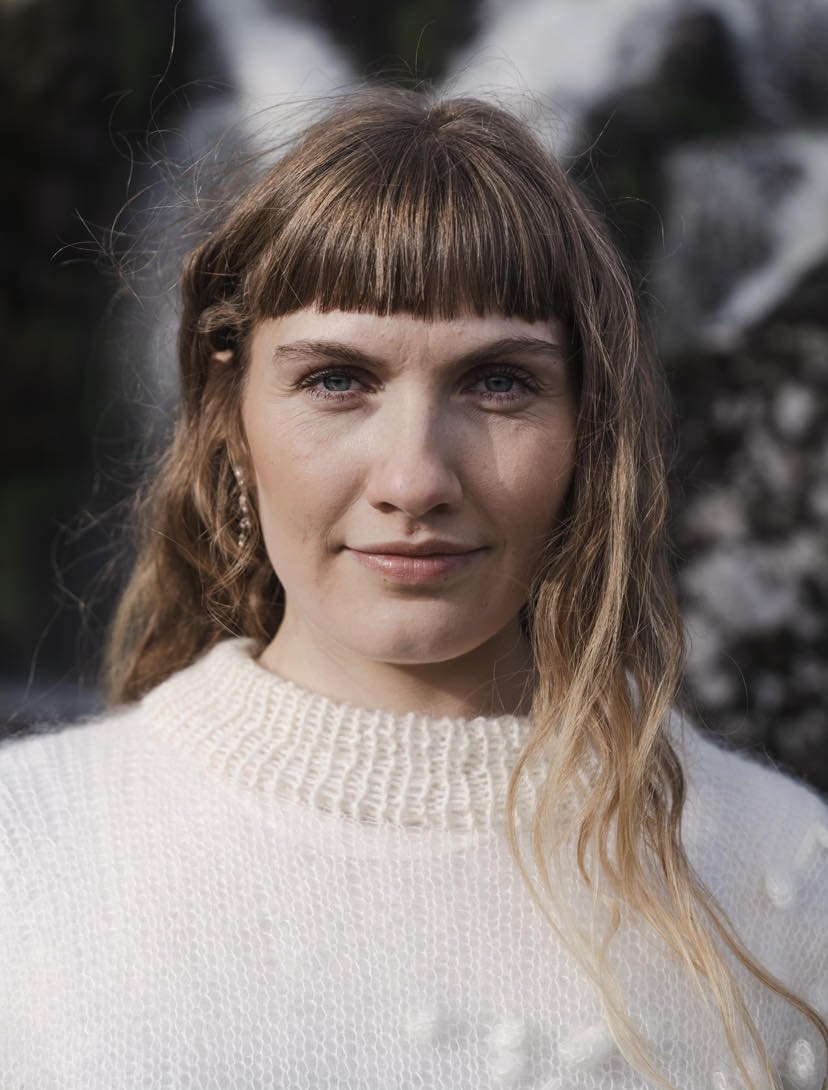 A woman with blonde hair and bangs, wearing a white knit sweater, looks directly at the camera with a neutral expression, outdoors with a blurred natural background.