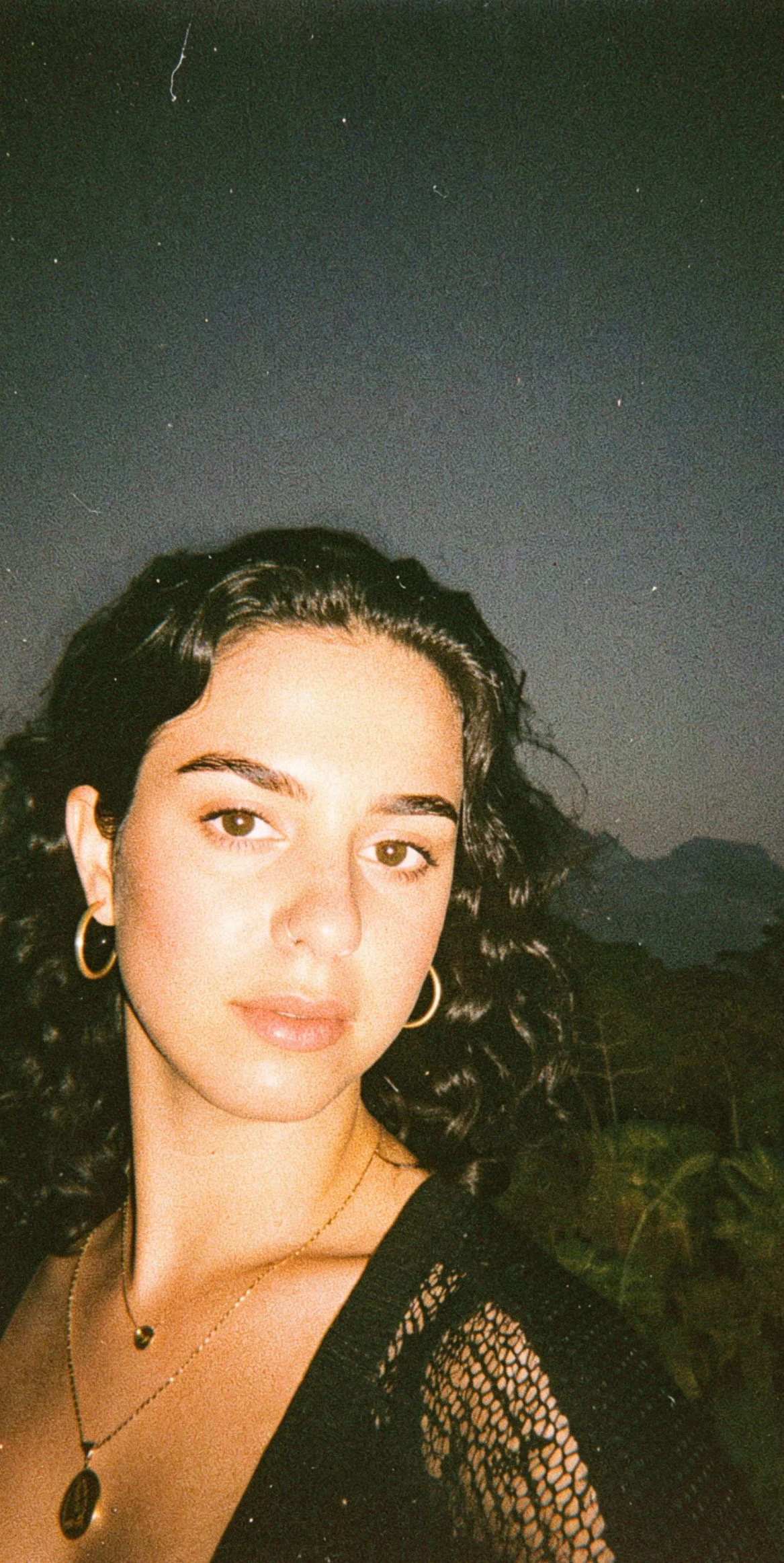A woman with dark curly hair, gold hoop earrings, and layered necklaces, posing outdoors at night with a dark sky and distant mountains in the background.