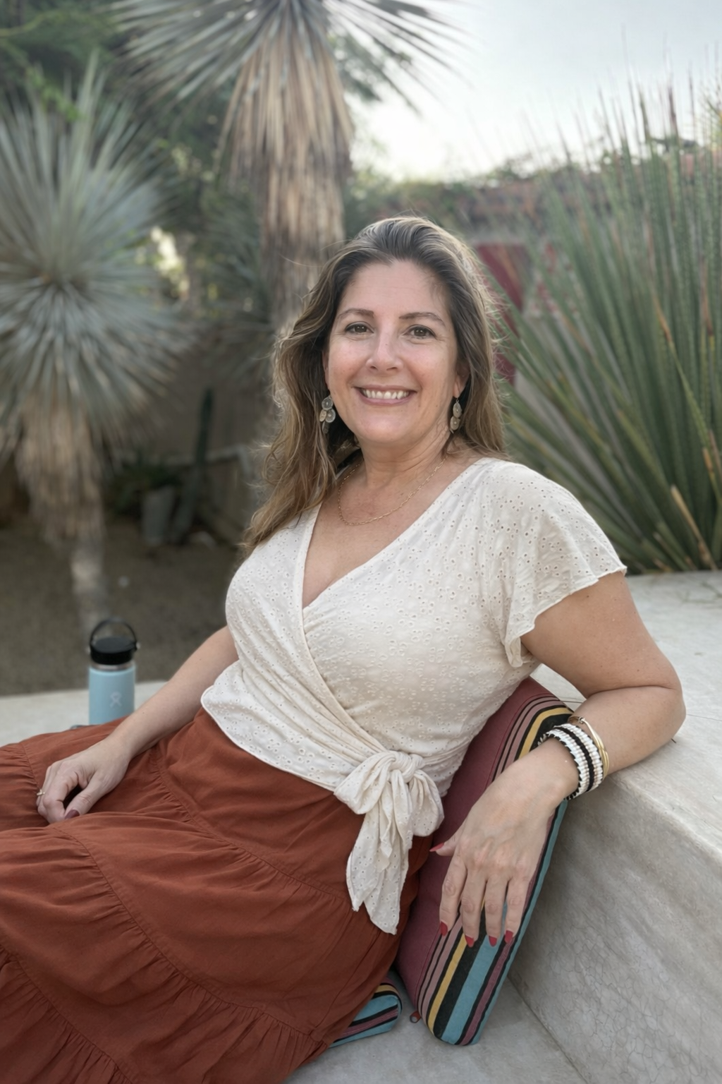 A smiling woman sitting outdoors near desert plants, wearing a cream-colored top and rust-colored skirt, with jewelry and a water bottle nearby.