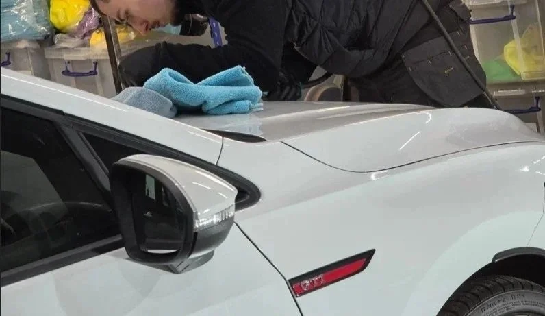 Person cleaning the hood of a white vehicle with a blue cloth in a garage or workshop.