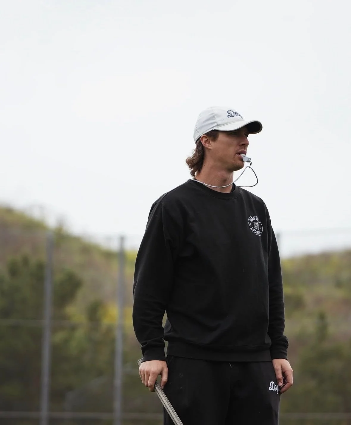 A young man with long brown hair wearing a black sweatshirt with a small logo and black pants, holding a golf club, outdoors on a cloudy day.