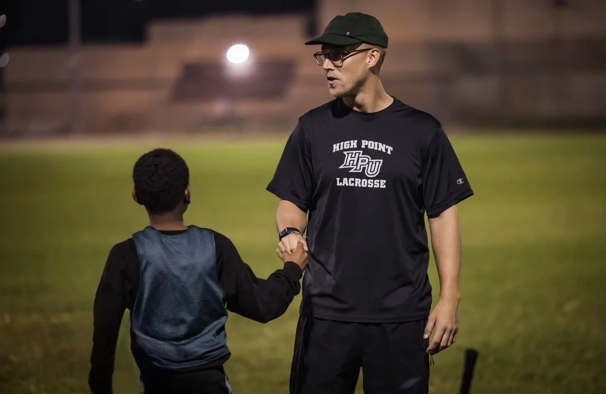 A man wearing glasses, a black cap, and a black High Point lacrosse T-shirt shakes hands with a young boy wearing a sports vest on a grassy field at night.