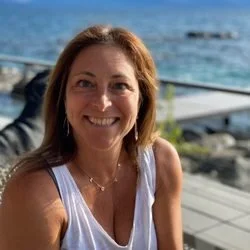 A woman smiling outdoors near the water with boats and rocks in the background.