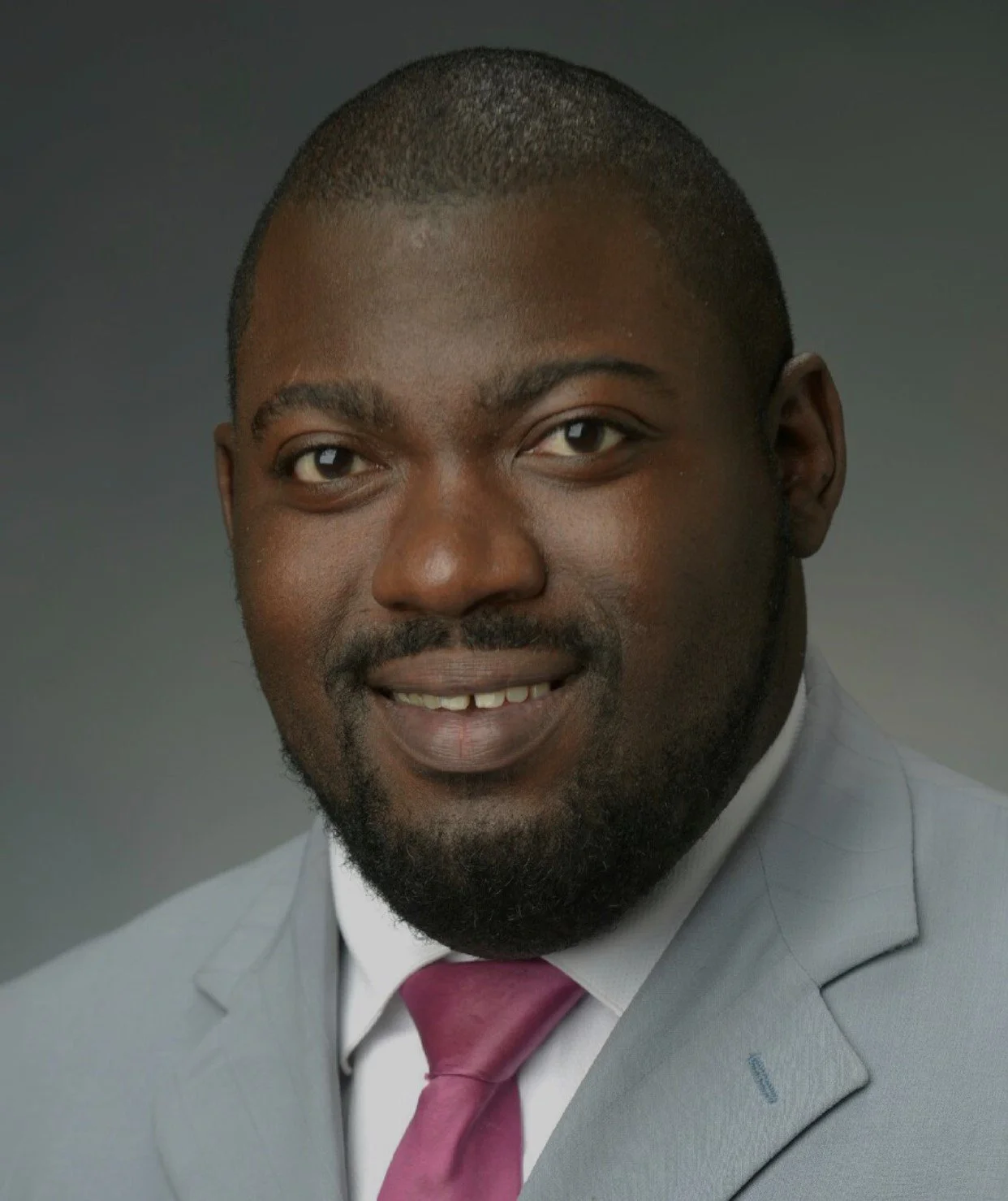 Headshot of a smiling African American man in a light gray suit, white shirt, and maroon tie, against a dark gray background.