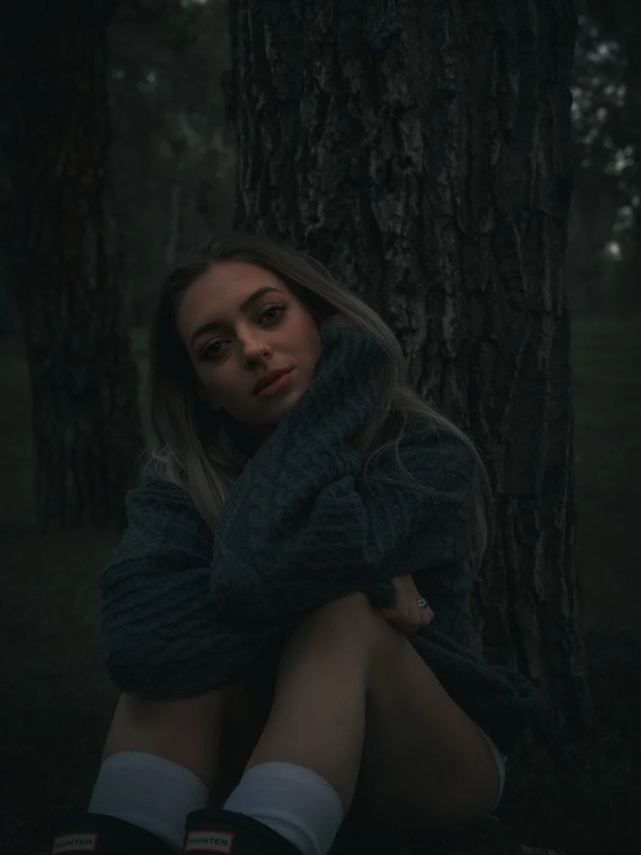 A young woman sitting outdoors near a large tree, wearing a dark sweater and white socks, with a contemplative expression.