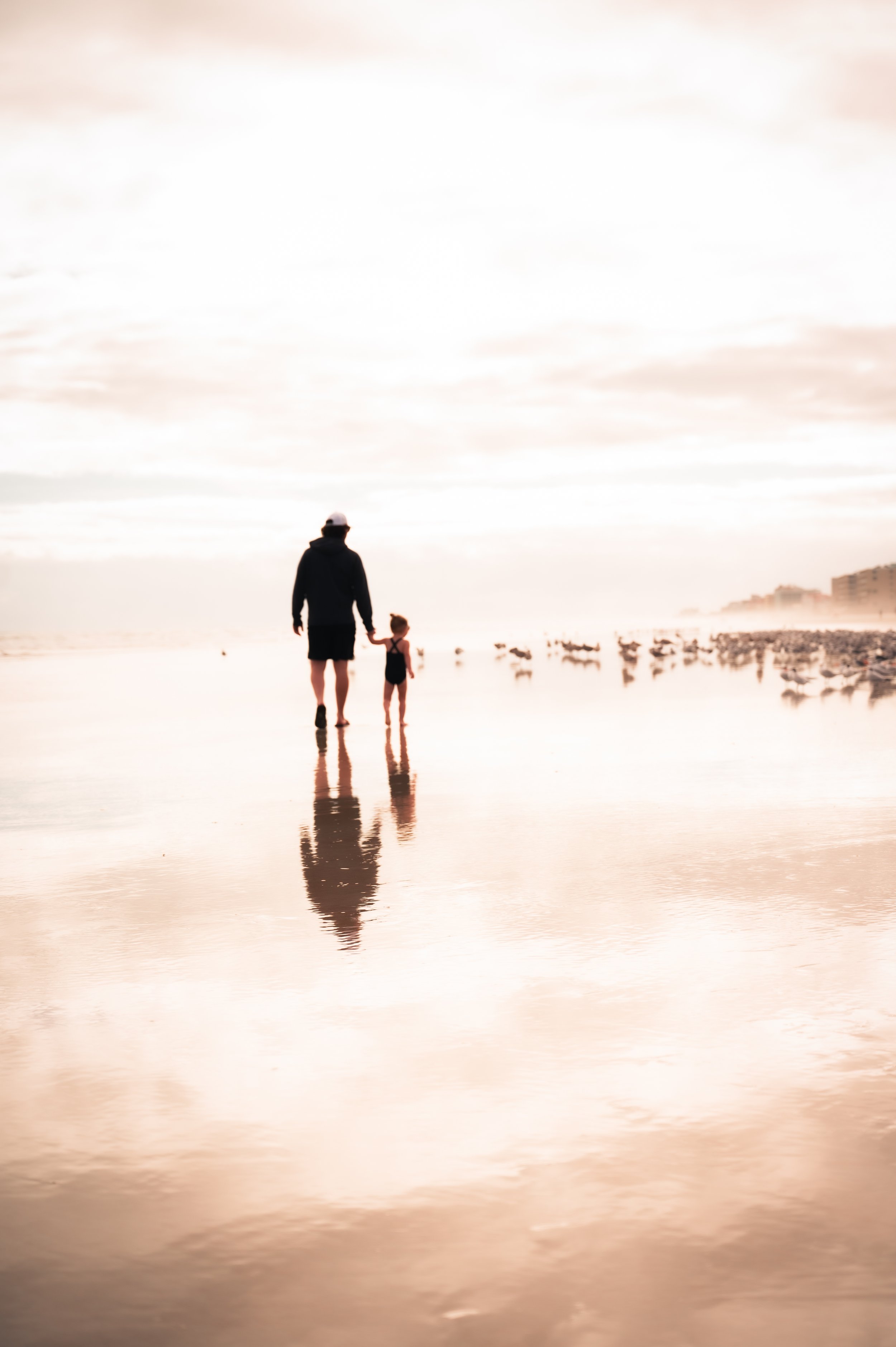 A person holding hands with a young girl walking along a beach shore during sunset, with their reflections visible in the shallow water.