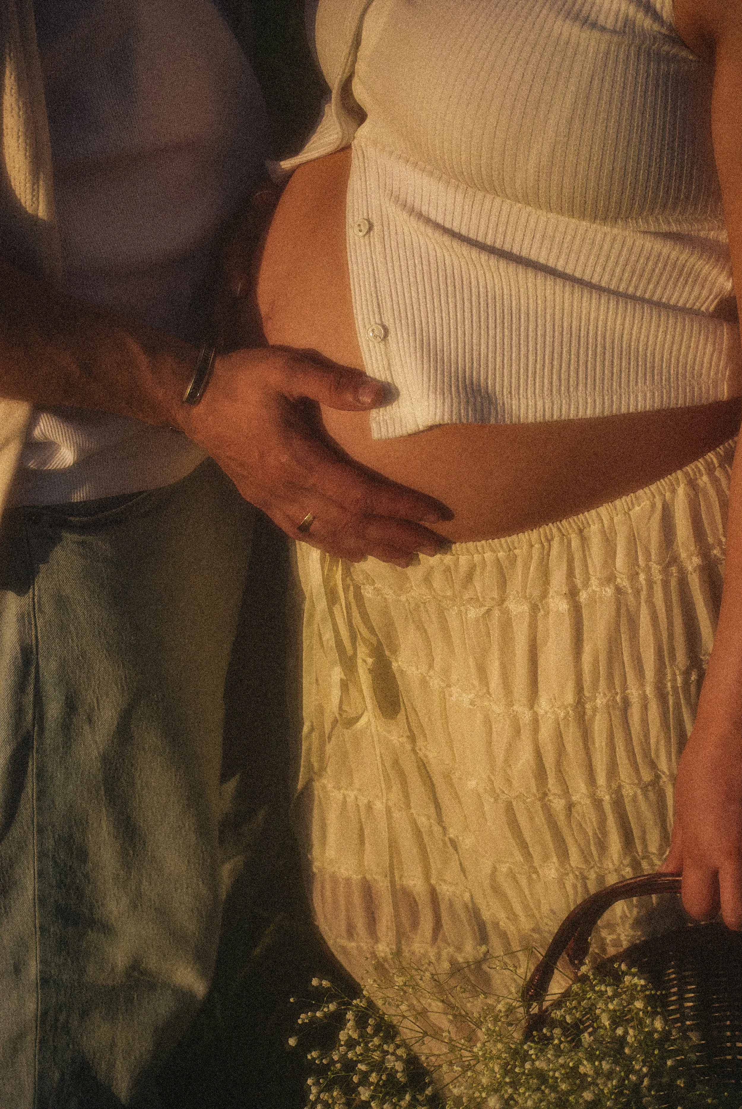 A person sitting on a bed, wearing a striped shirt and beige skirt, with their hand resting on their bare belly, with another person leaning over them, touching their stomach.