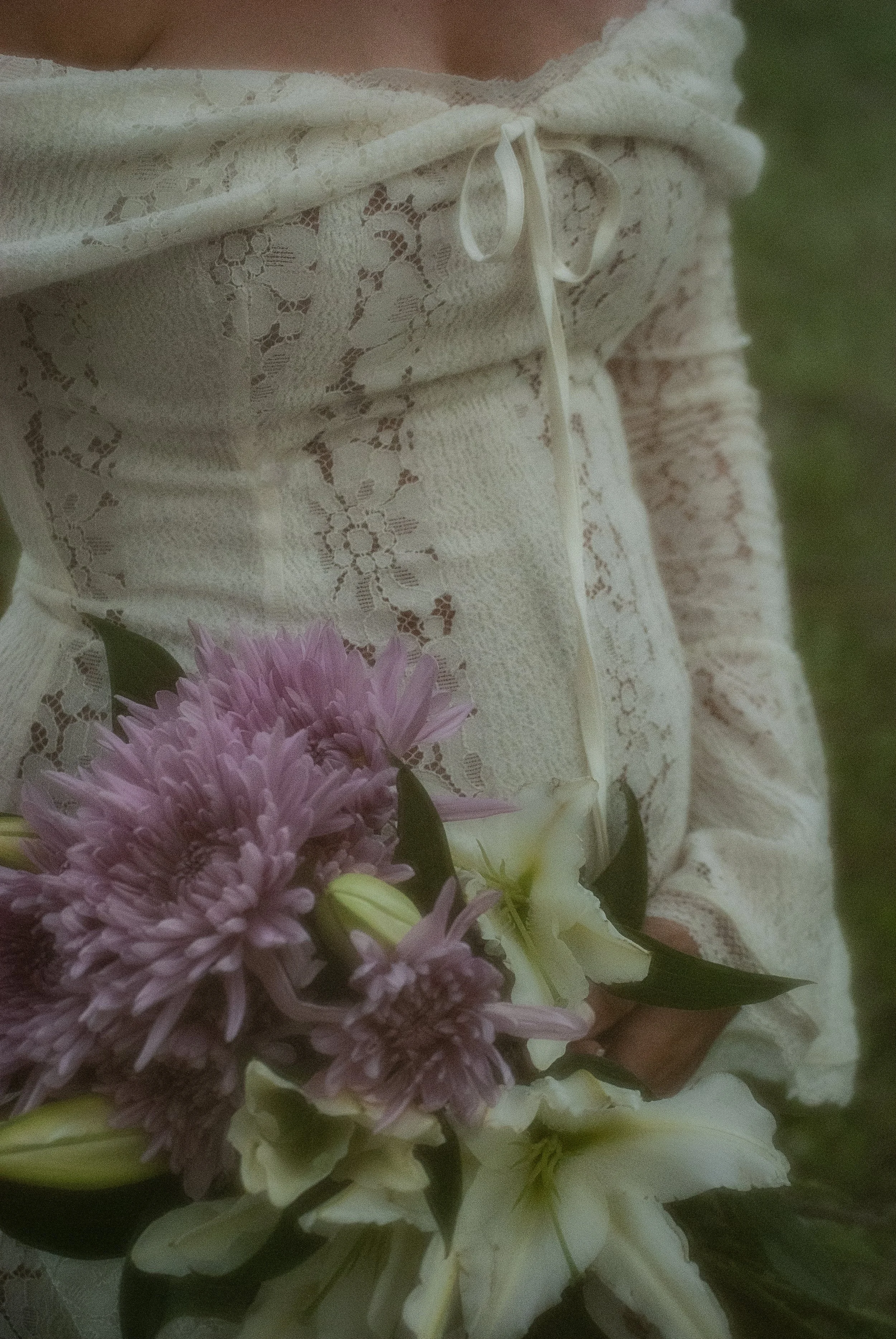 A person in a cream-colored lace dress holding a bouquet of purple and white flowers, with a blurred green background.