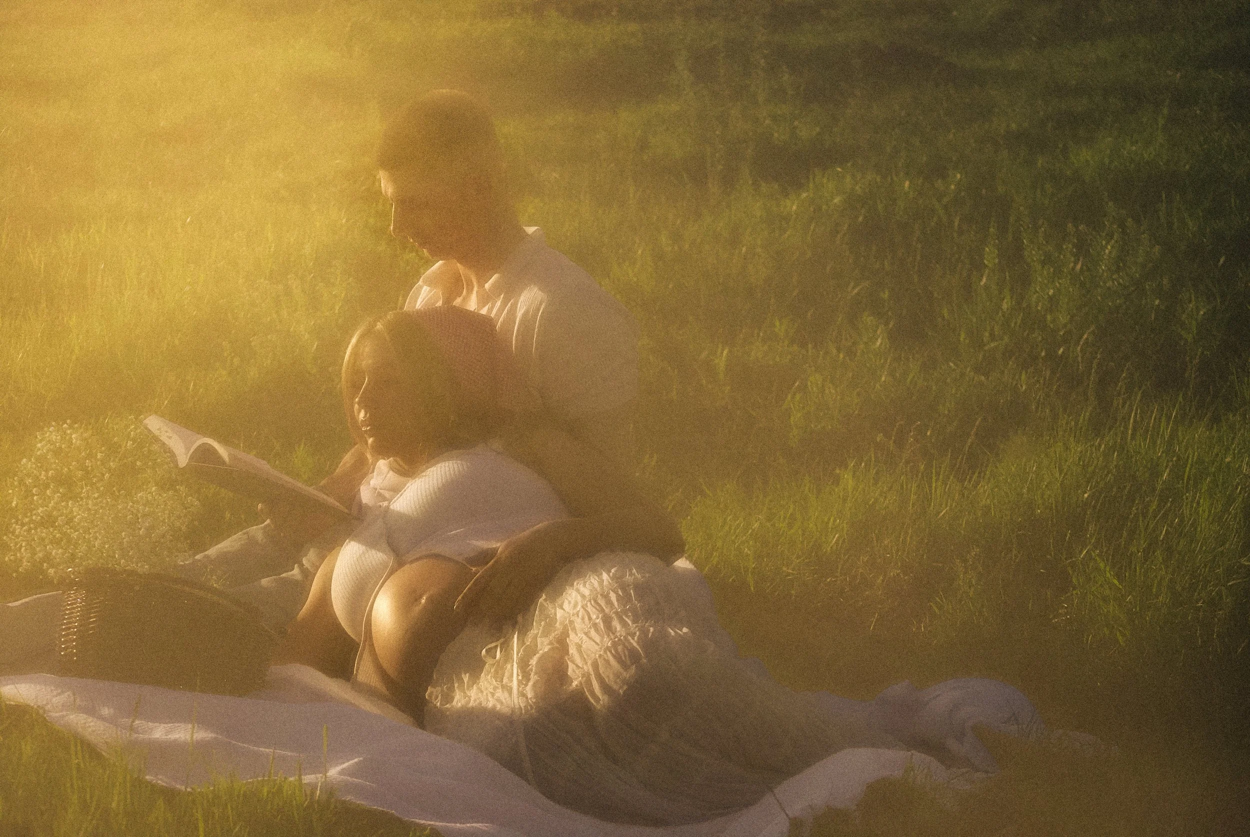A couple sitting on a blanket in a grassy field during sunset, reading a book together.