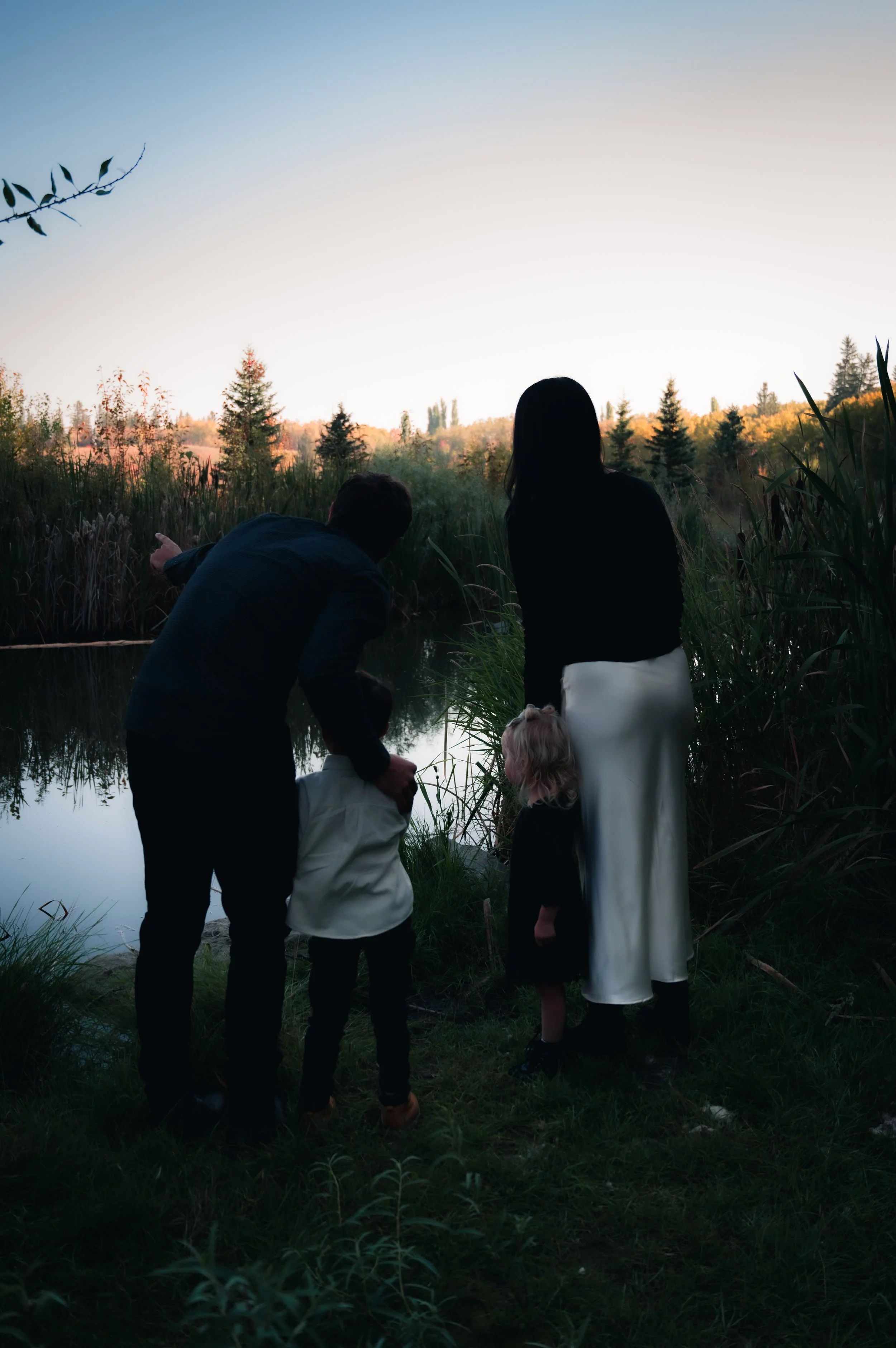 A family of five, including two adults and three children, standing by a river and looking at the water during sunset, surrounded by tall grass and trees.