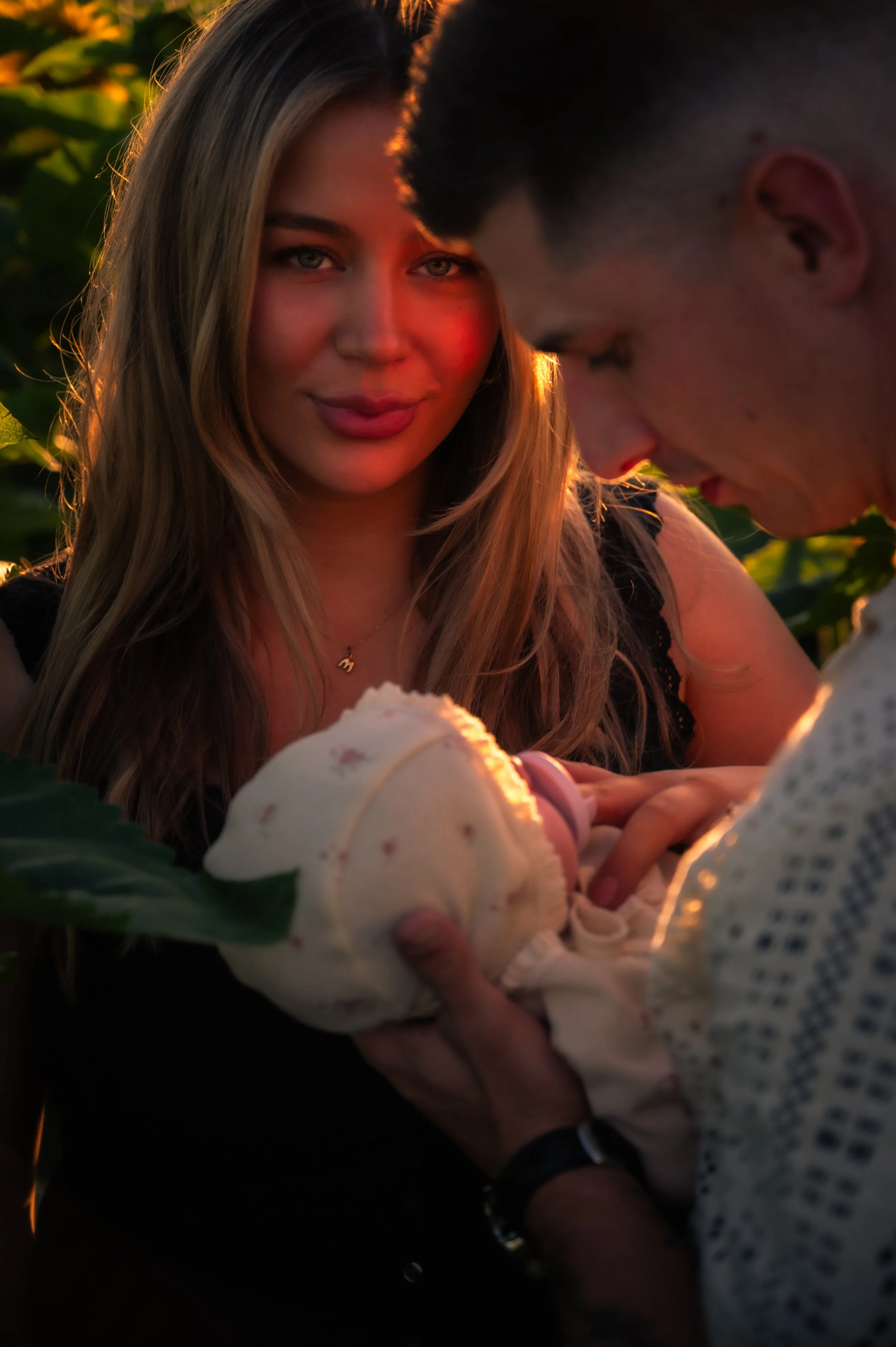 A woman receiving a baby from a man during sunset in a field of green plants.