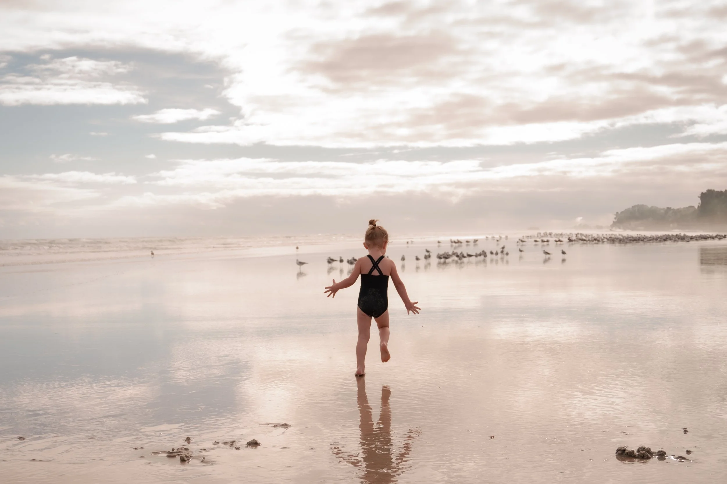 A young girl in a black swimsuit playing on a beach with reflections of the cloudy sky on wet sand, seagulls in the background, during a calm and overcast day.