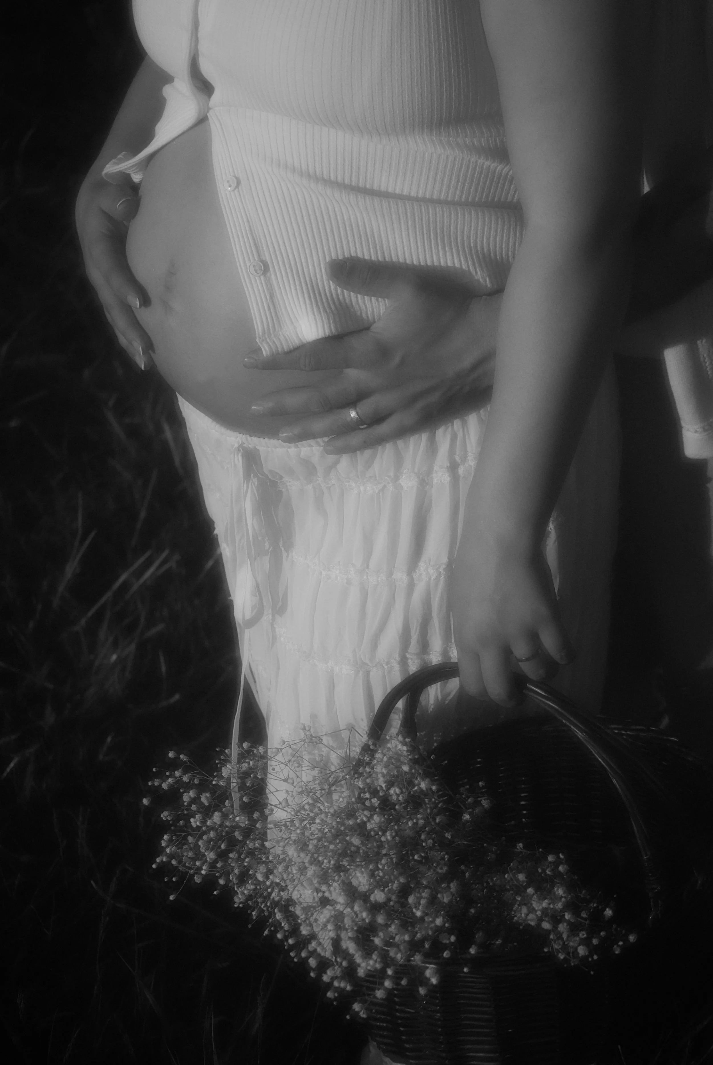 A pregnant woman lying on grass, wearing a striped blouse and white skirt, holding a basket of small flowers and with her hand resting on her belly.