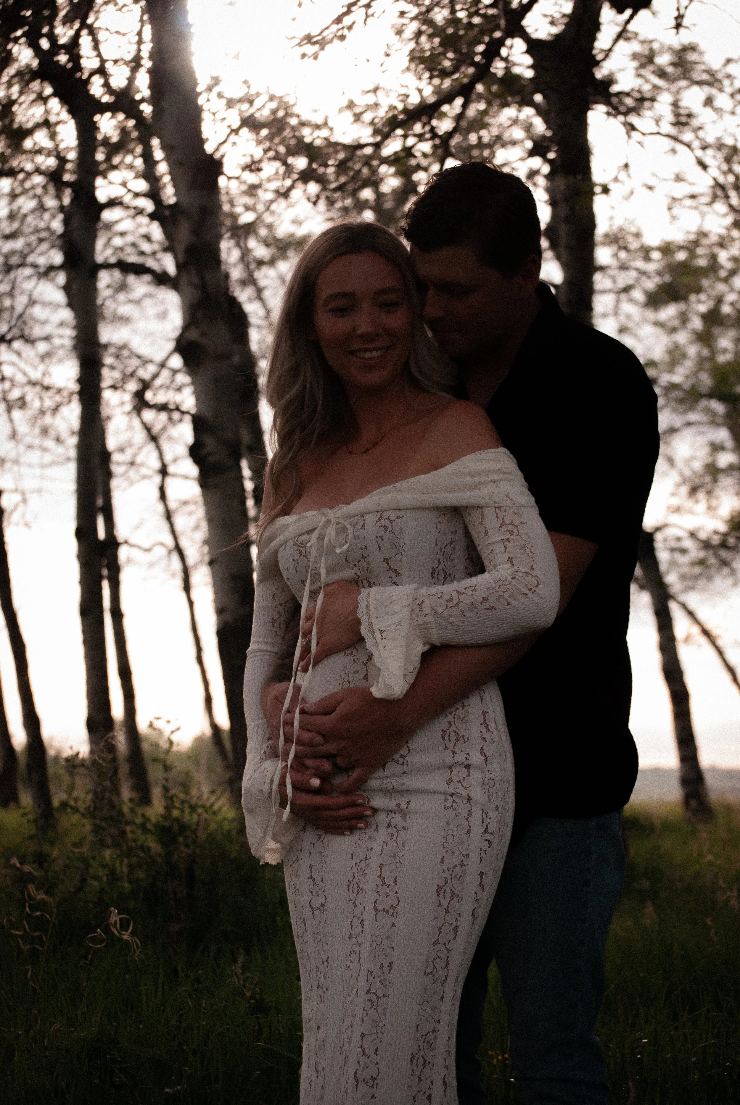 A couple, a woman in a white lace dress and a man in a black shirt, sharing a tender moment outdoors among trees during sunset.