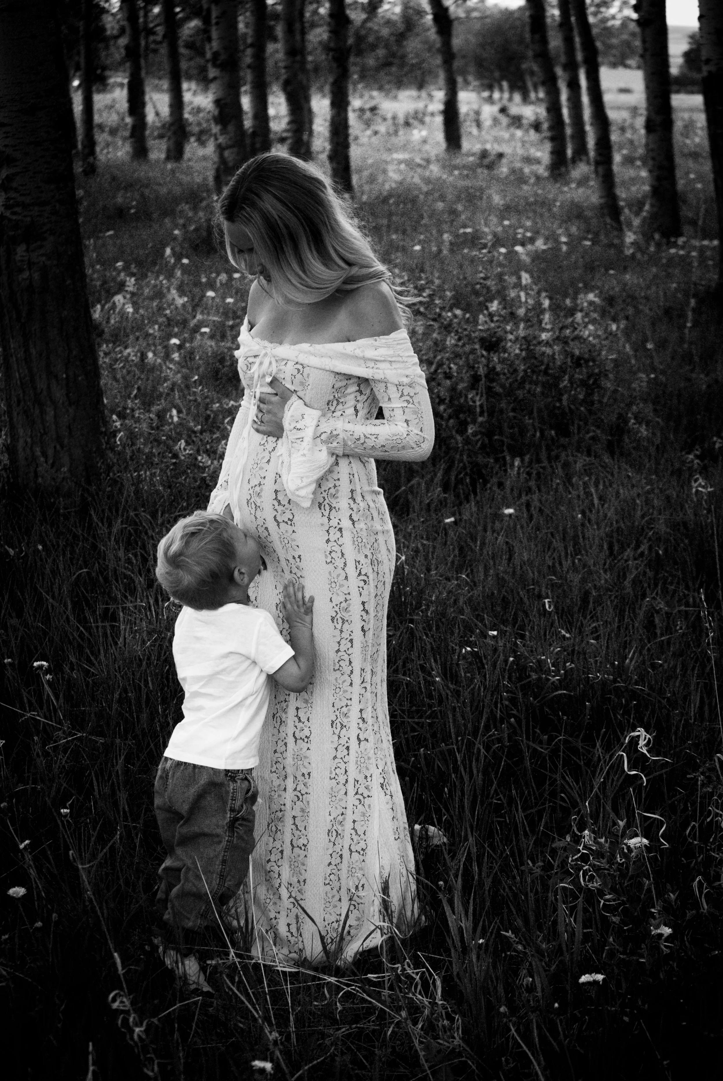 Black and white photo of a woman in a long, lace dress standing in a grassy field surrounded by trees, with a young boy touching her dress and looking up at her.