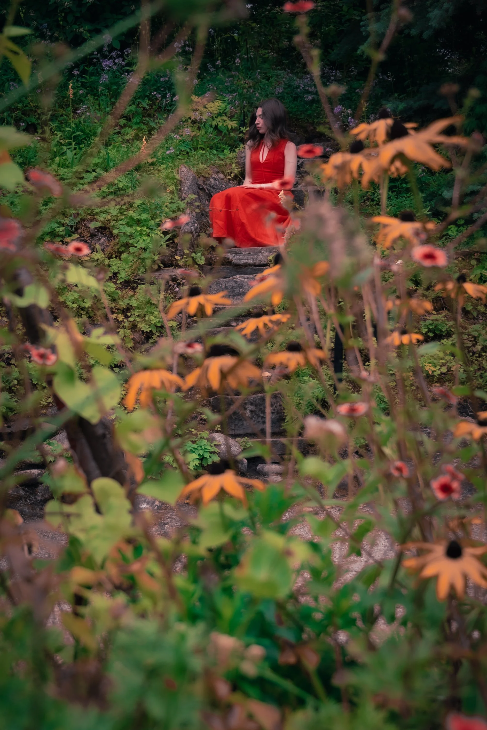 A woman in a red dress sitting on a rock surrounded by lush greenery and colorful flowers.