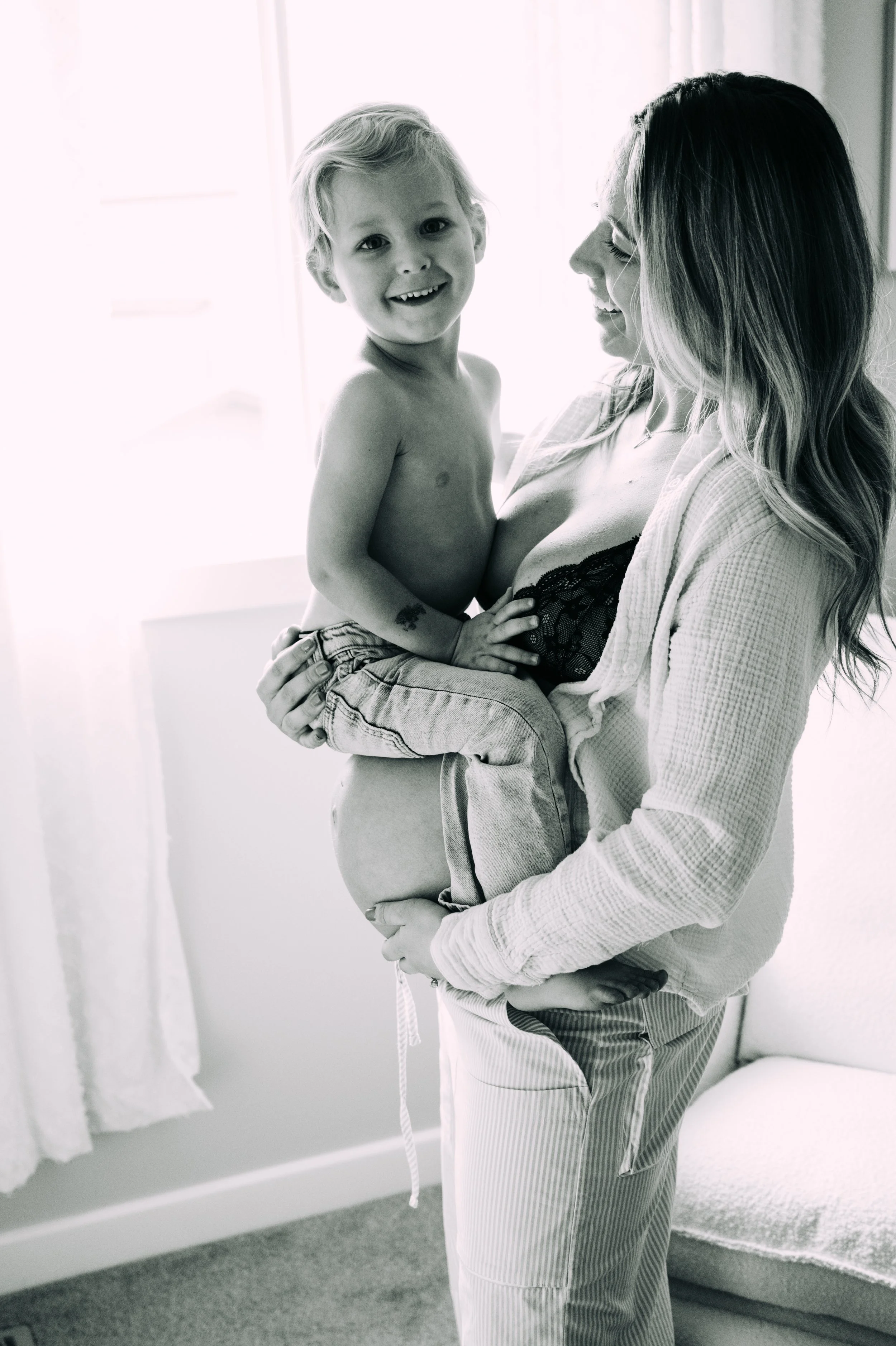 A woman holding a smiling young boy in a bright room with curtains and a chair.