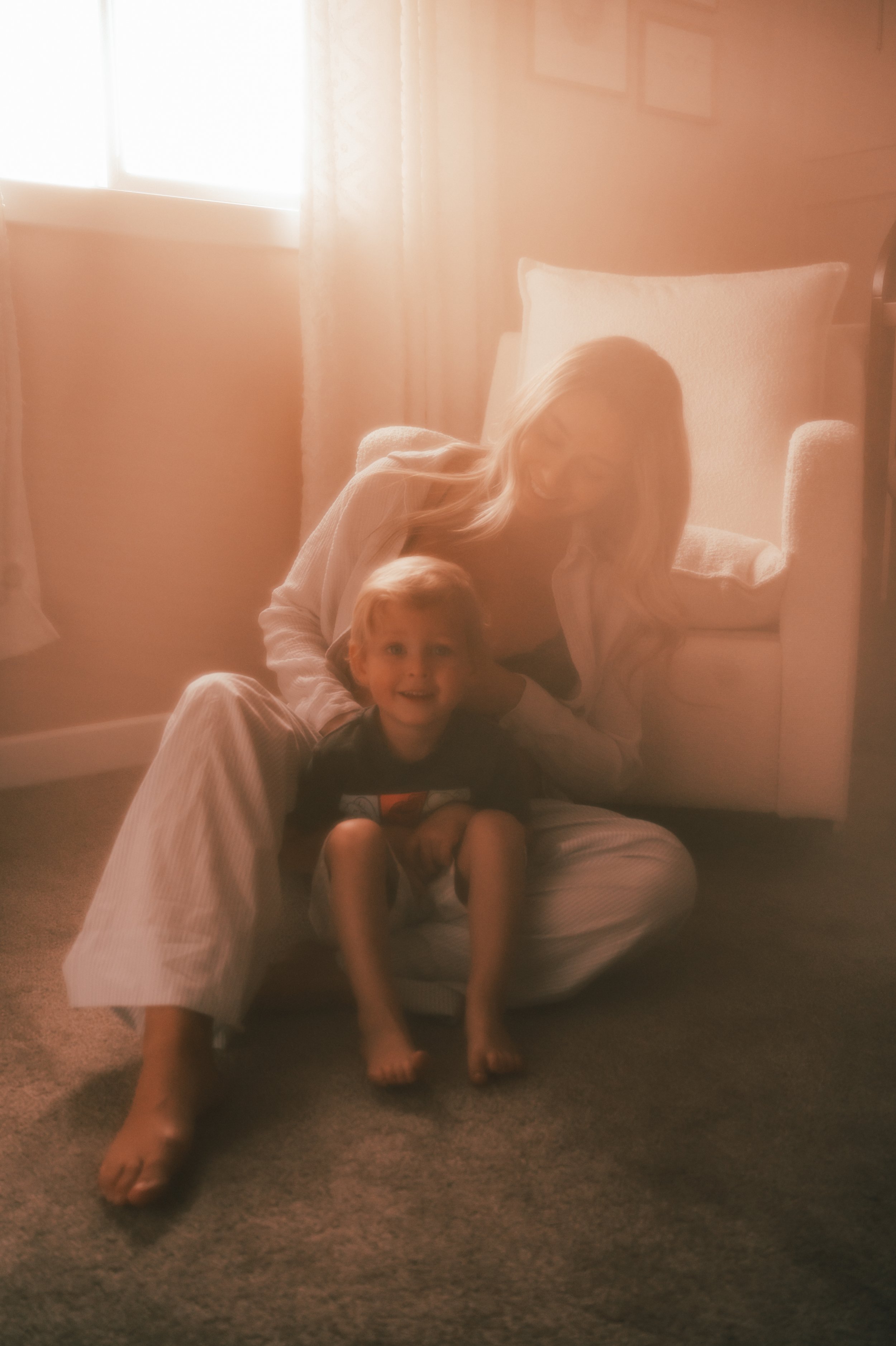A woman and a young boy sitting on a carpeted floor near a window with sunlight streaming in. The woman is smiling and leaning on a light-colored armchair, while the boy is sitting cross-legged and smiling at the camera.