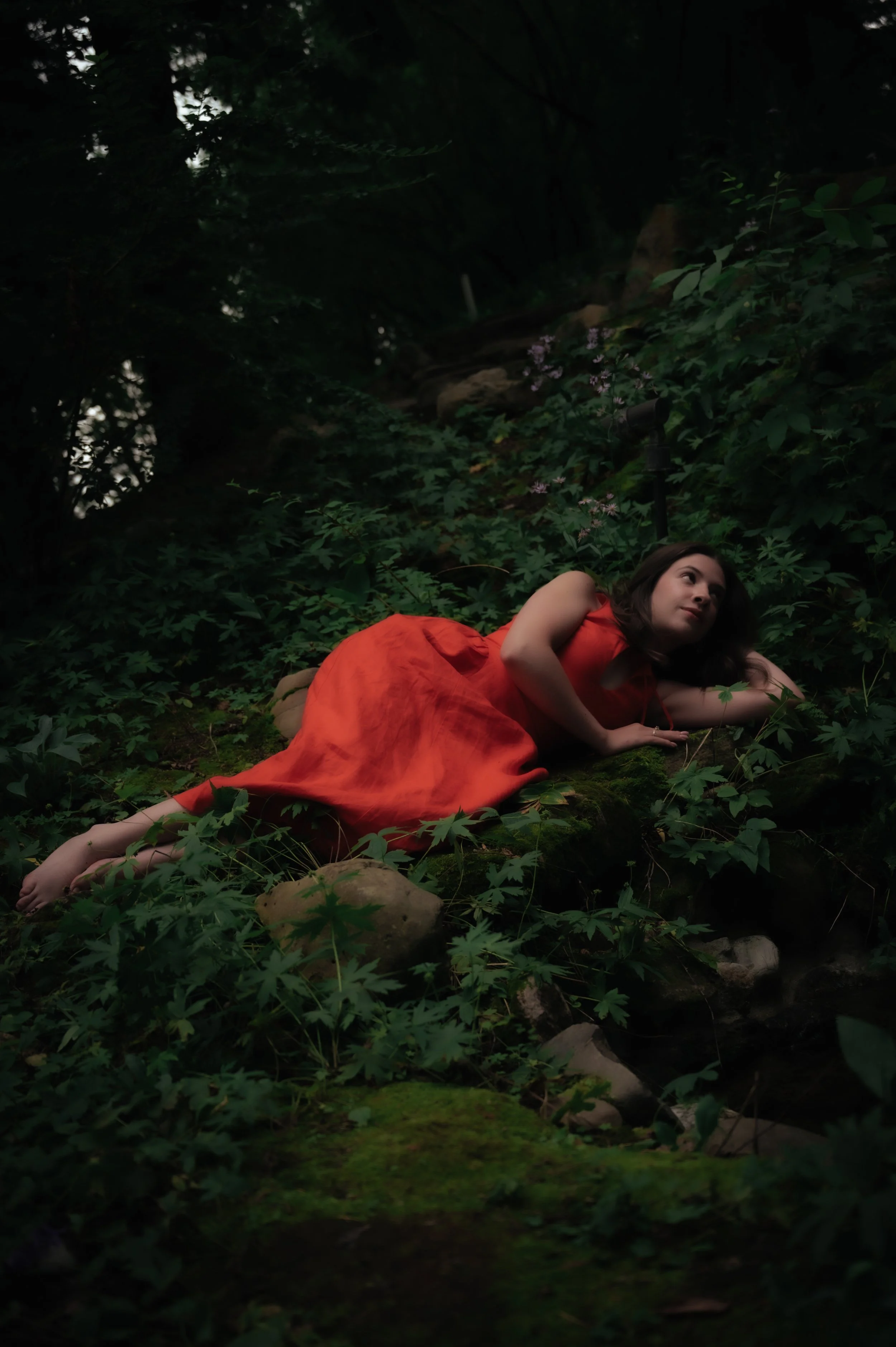 A woman in a red dress lying on her side in a lush, green forest with rocks and moss, looking contemplative.