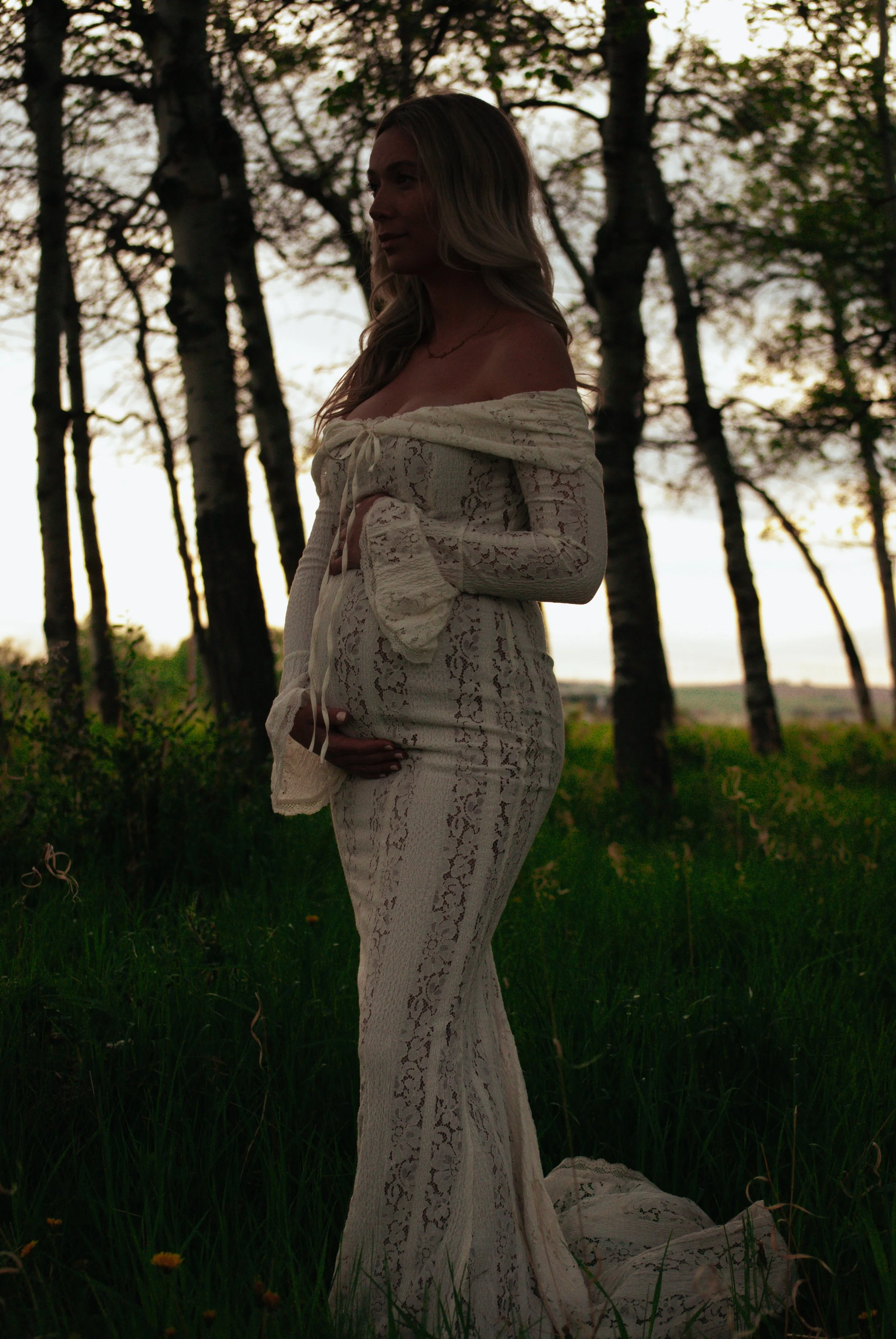 A woman in a long, off-shoulder cream lace dress standing outdoors in a grassy area with trees and a sunset in the background.