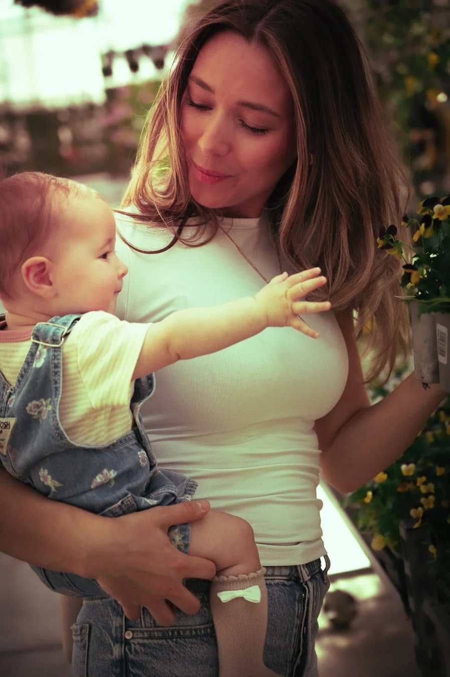 A woman with long brown hair holds a baby girl while shopping for flowers in a greenhouse. The baby is reaching out towards a flower plant.