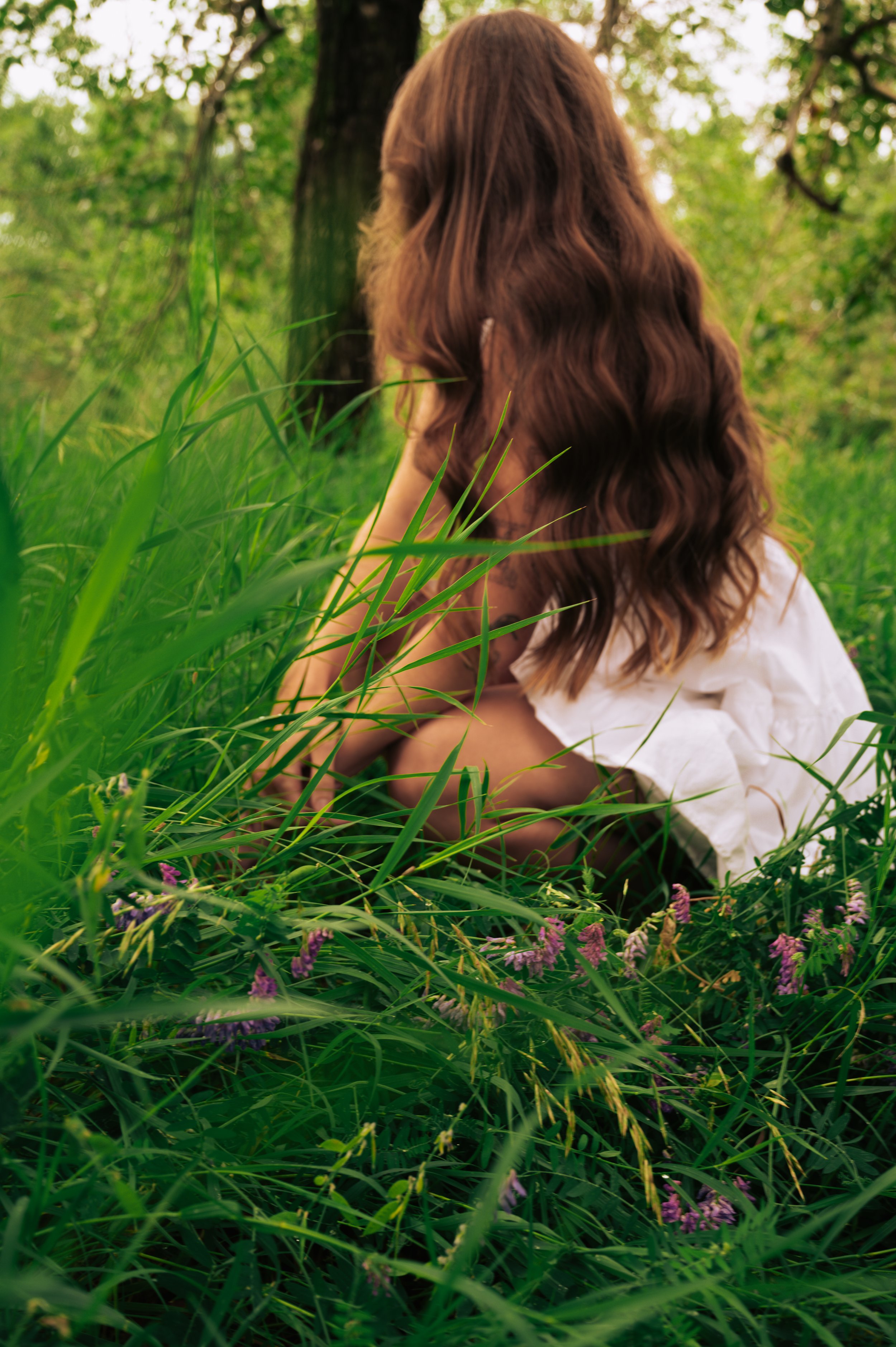 A woman with long, wavy brown hair crouching in tall green grass and plants in a forested area.