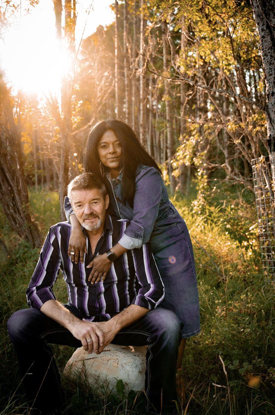 A woman with long black hair and dark skin standing behind an older man with gray hair and light skin, sitting on a rock in a wooded area during sunset, both looking at the camera.
