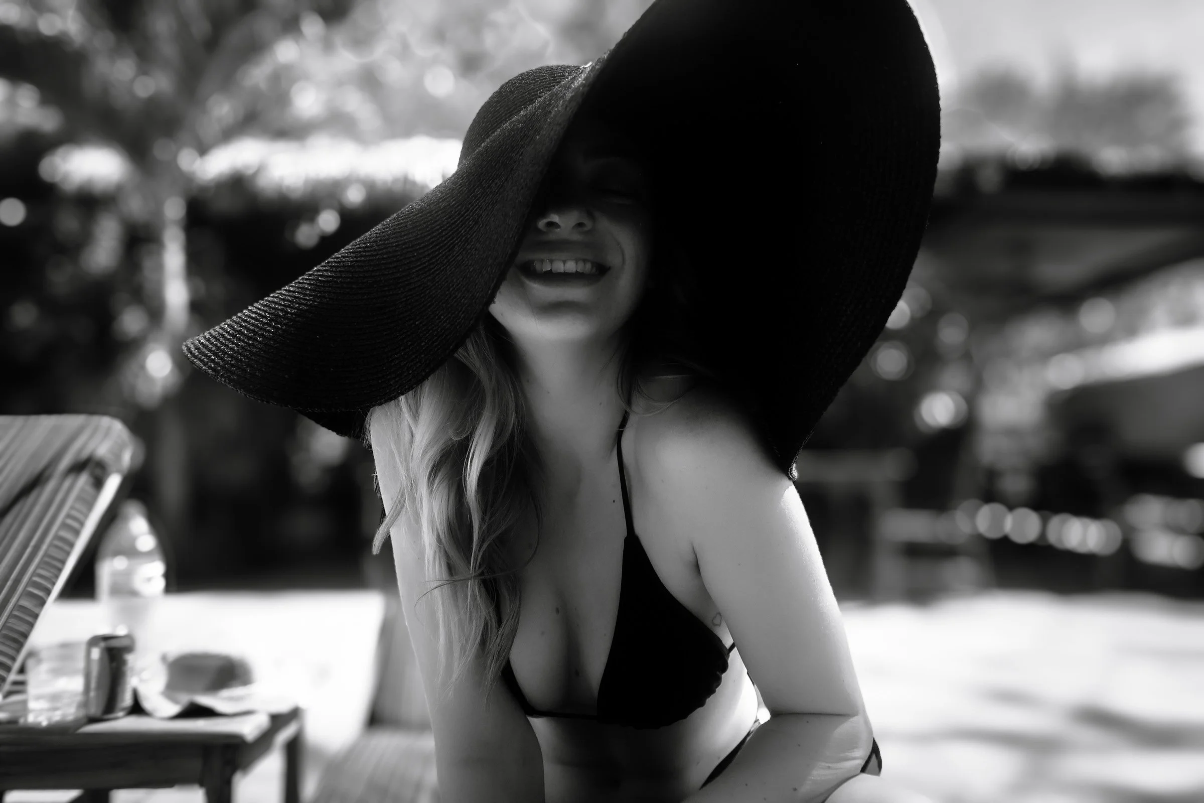 A woman wearing a large black sunhat and a bikini top, smiling on a beach.